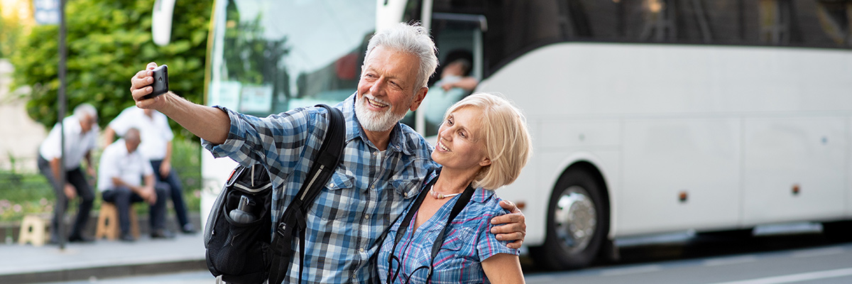 An older couple taking a selfie in front of a tour bus and smiling